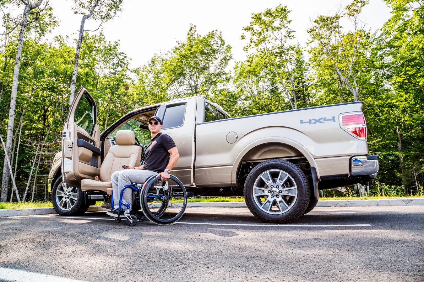 A man is sittting in a wheelchair next to the OEM seat attached to the LINK turning seat base deployed on the driver's side of a pickup truck.